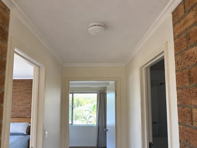 Ceiling-mounted smoke alarm in a hallway with brick walls and doorways in Currumbin Waters